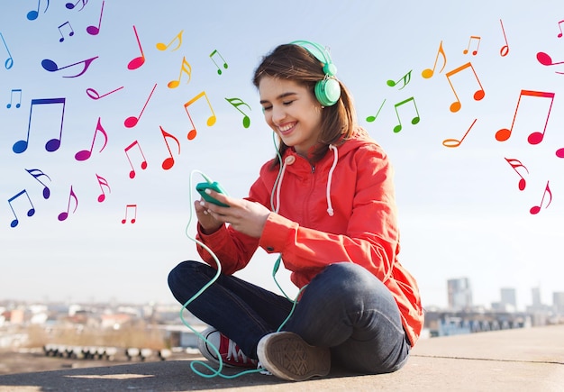 conceito de tecnologia, estilo de vida e pessoas - jovem sorridente ou adolescente com smartphone e fones de ouvido ouvindo música ao ar livre sobre fundo colorido de notas musicais