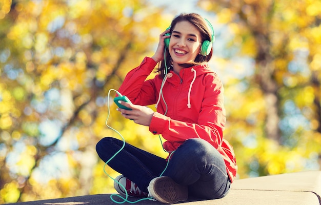 conceito de tecnologia, estilo de vida e pessoas - jovem sorridente ou adolescente com smartphone e fones de ouvido ouvindo música ao ar livre sobre o fundo do parque outono