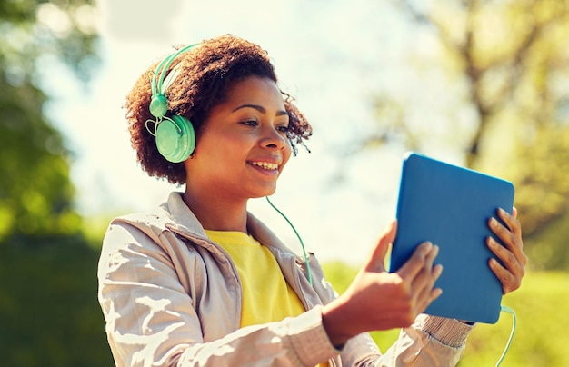 conceito de tecnologia, estilo de vida e pessoas - sorrindo jovem afro-americana ou adolescente com computador tablet pc e fones de ouvido ouvindo música ou assistindo vídeo no parque de verão
