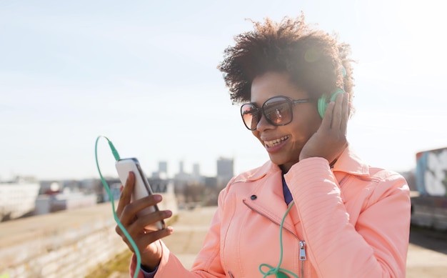 Conceito de tecnologia, estilo de vida e pessoas - sorrindo jovem afro-americana ou adolescente com smartphone e fones de ouvido ouvindo música ao ar livre