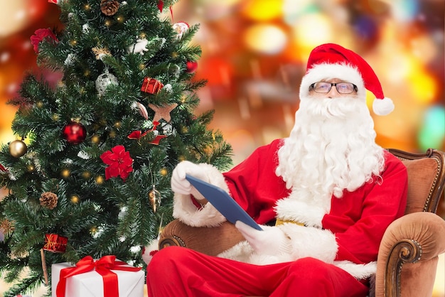 conceito de tecnologia, feriados e pessoas - homem fantasiado de papai noel com computador tablet pc, presentes e árvore de natal sentado na poltrona sobre fundo de luzes vermelhas