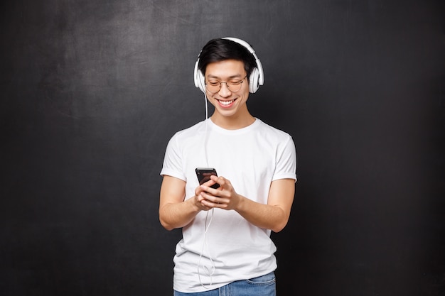 Conceito de tecnologia, gadgets e pessoas. Feliz sorridente jovem asiático bonito de camiseta, ouvir música em fones de ouvido, escolher lista de reprodução no telefone móvel, amigo de mensagens de texto