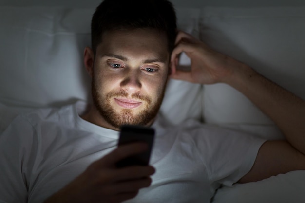 Foto conceito de tecnologia, internet, comunicação e pessoas - jovem enviando mensagens de texto no smartphone na cama em casa à noite