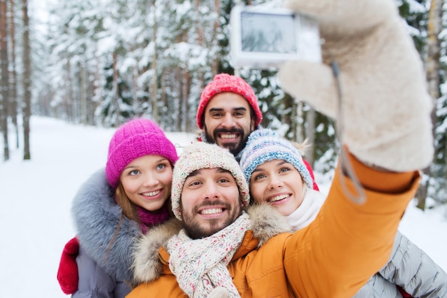 conceito de tecnologia, temporada, amizade e pessoas - grupo de homens e mulheres sorridentes tomando selfie com câmera digital na floresta de inverno