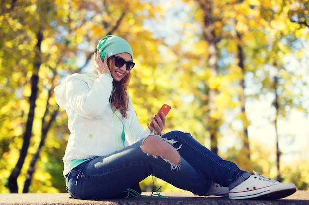 Foto conceito de tecnologia, temporada e pessoas - jovem sorridente ou adolescente com smartphone e fones de ouvido ouvindo música sobre o fundo do parque outono