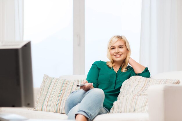 conceito de televisão, lazer e pessoas - mulher sorridente sentada no sofá com controle remoto e assistindo tv em casa