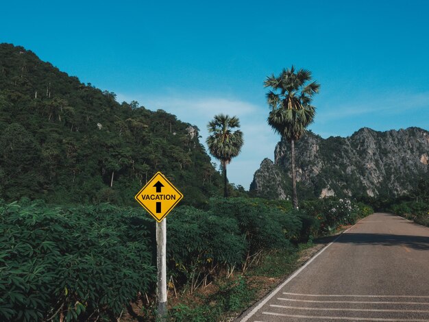 Conceito de tempo de férias. Palavras "férias" e uma seta direta na placa amarela em pé perto da estrada rural cercam com árvores verdes e montanhas no fundo do céu azul.