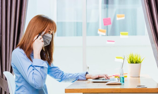 Conceito de trabalho em casa, jovem empresária asiática usando máscara facial quarentena protetora doença coronavírus pandêmico ou COVID-19 ela trabalhando na mesa com laptop no escritório doméstico