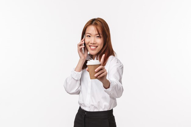 Conceito de trabalho, pessoas e estilo de vida de escritório. Alegre jovem asiática boba, sugerindo-lhe a xícara de café enquanto fala no telefone, sorrindo câmera alegre, ter uma conversa,