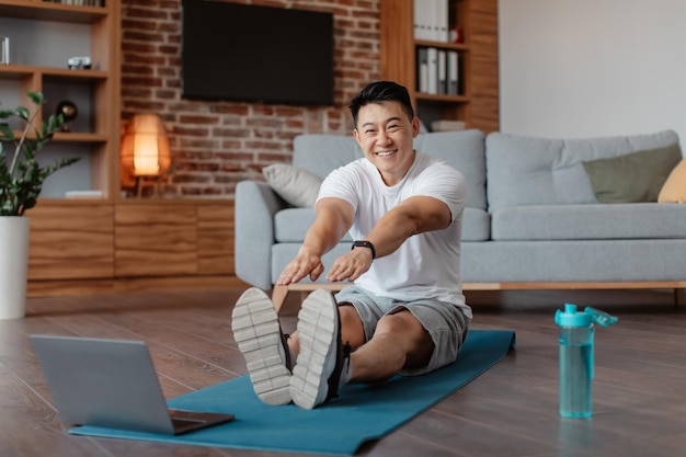 Conceito de treinamento em casa Homem coreano positivo esticando os músculos da perna na frente do laptop assistindo a tutoriais em vídeo on-line