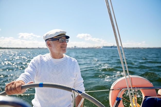 conceito de vela, idade, turismo, viagens e pessoas - feliz homem sênior com chapéu de capitão no volante e navegando em veleiro ou iate flutuando no mar