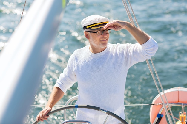 conceito de vela, idade, turismo, viagens e pessoas - feliz homem sênior com chapéu de capitão no volante e navegando em veleiro ou iate flutuando no mar