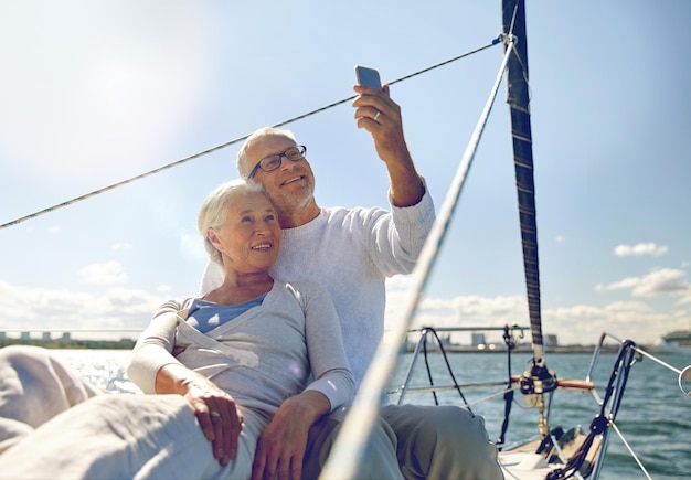 conceito de vela, tecnologia, turismo, viagens e pessoas - casal sênior feliz tomando selfie com smartphone em barco a vela ou convés de iate flutuando no mar