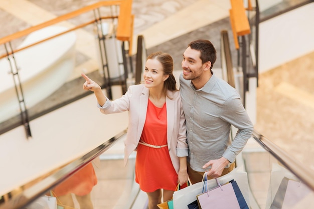 conceito de venda, consumismo e pessoas - casal jovem feliz com sacolas de compras subindo na escada rolante e apontando o dedo no shopping