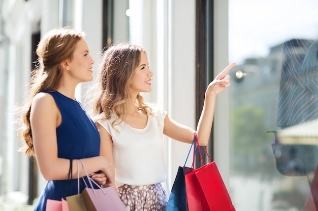 conceito de venda, consumismo e pessoas - mulheres jovens felizes com sacolas de compras apontando o dedo para a vitrine na cidade