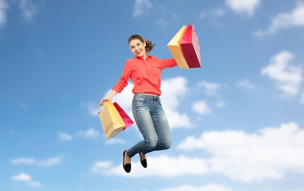 conceito de venda, movimento e pessoas - jovem sorridente com sacolas de compras pulando no ar sobre fundo azul do céu e nuvens