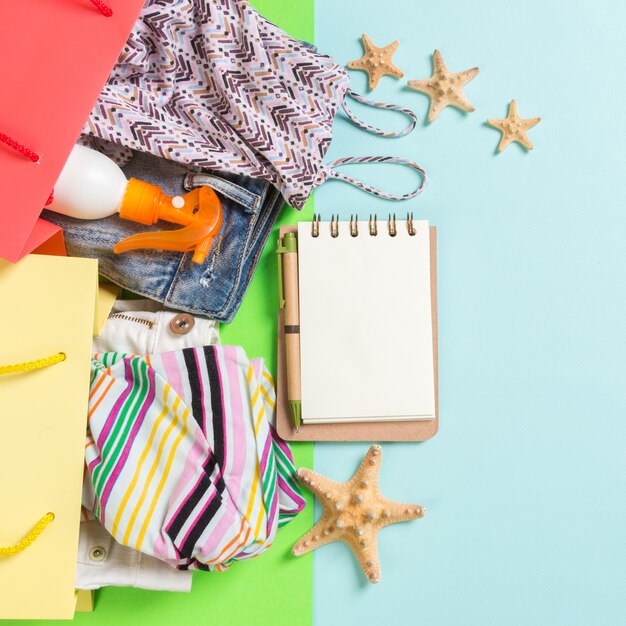 Conceito de verão de sacolas coloridas cheias de roupas. Sacos de presente com camiseta, bermuda jeans, caderno e frasco de protetor solar. Conceito de lista de desejos de verão. Espaço da cópia da vista superior sobre fundo azul.