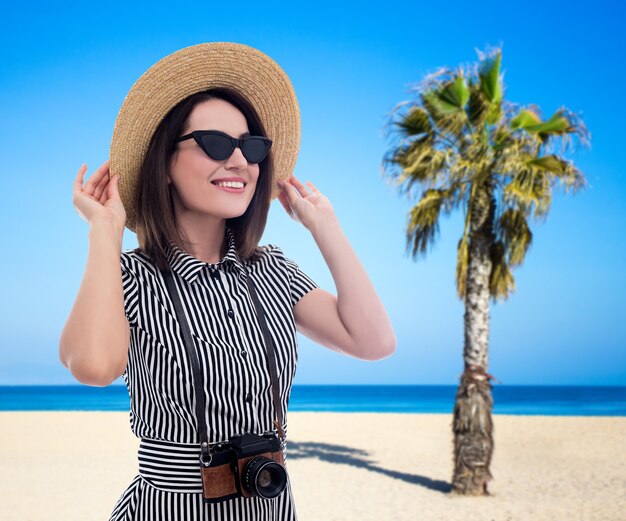 Conceito de verão e viagens - jovem mulher bonita com câmera caminhando na praia com palmeiras