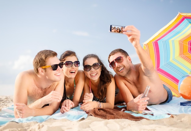 conceito de verão, férias, férias, tecnologia e felicidade - grupo de pessoas sorridentes em óculos de sol tirando foto com smartphone na praia