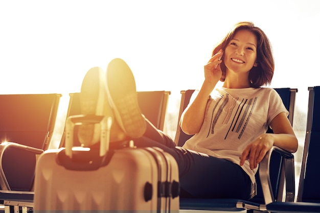 Conceito de viagem aérea com jovem casual sentada com mala de bagagem de mão Mulher do aeroporto falando ao telefone no portão esperando no terminal