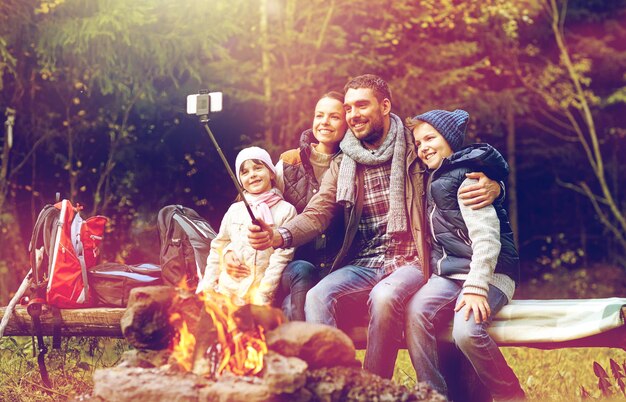 Foto conceito de viagem, turismo e caminhada - família feliz sentada em um banco e tirando uma foto com um smartphone em um selfie stick em uma fogueira na floresta