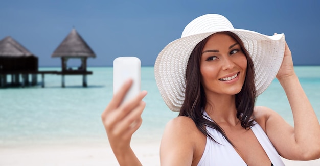 conceito de viagem, verão, tecnologia e pessoas - jovem sexy tirando selfie com smartphone sobre bangalô no fundo da praia