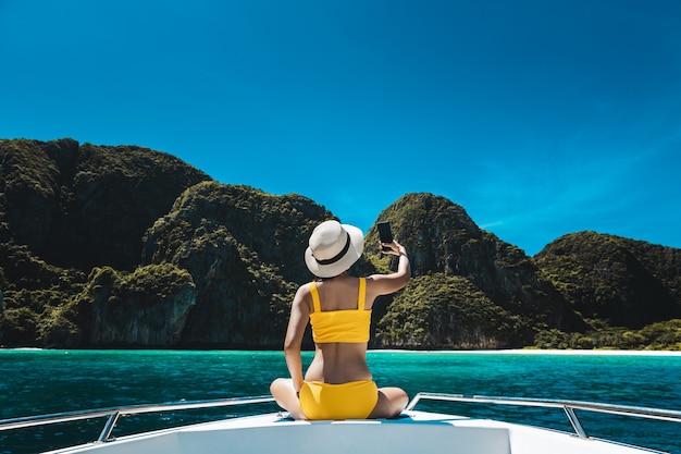 Conceito de viagens de férias de verão. Mulher asiática feliz viajante solo com biquíni e telefone celular relaxa e selfie em um barco na Baía Maya de Phuket, Tailândia