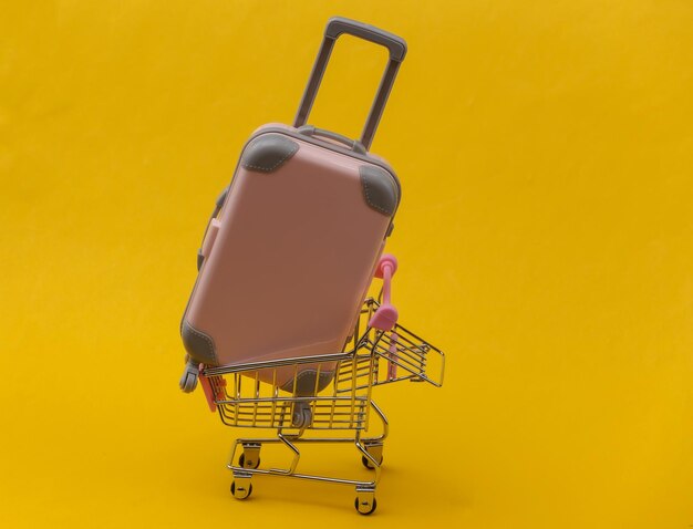 Foto conceito de viagens e compras. mini mala de viagem de plástico rosa e carrinho de supermercado em fundo amarelo. estilo minimalista