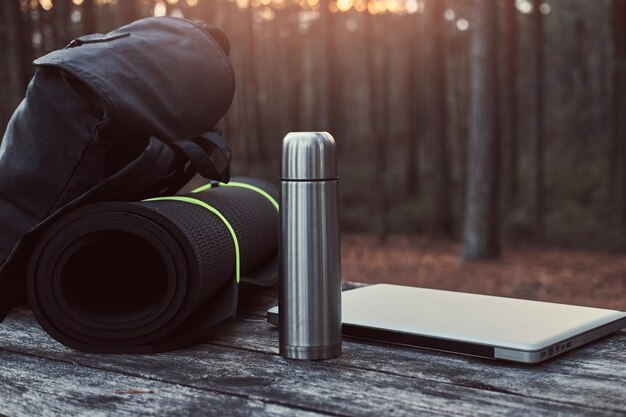 Conceito de viagens. Mochila, tapete turístico, laptop, smartphone e garrafa térmica em uma mesa de madeira em uma bela floresta.