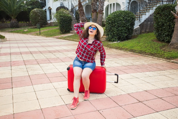 Conceito de viagens, turismo e pessoas. garota feliz sentada na mala vermelha de óculos e mantém o chapéu na cabeça.