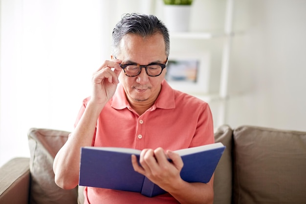 conceito de visão, lazer, literatura e pessoas - homem de óculos sentado no sofá e lendo livro em casa