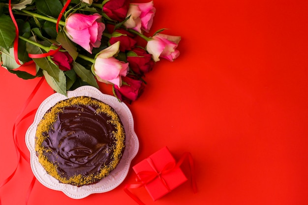 Conceito do dia da mulher ou dia dos namorados St. Rosas frescas e caixa de presente, sobre um fundo vermelho e um bolo de chocolate. Vista do topo. Copie o espaço