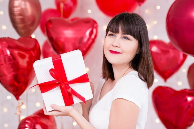 Conceito do dia dos namorados - mulher sonhadora feliz com caixa de presente sobre fundo vermelho de balões em forma de coração