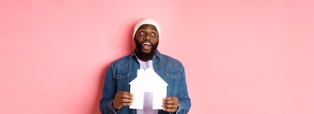 Conceito imobiliário homem afro-americano bonito sonhando com casa segurando modelo de casa e olhando