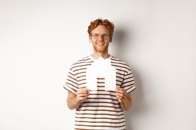 Conceito imobiliário. homem ruivo sorridente de óculos segurando a casa de papel recortado e olhando para a câmera, economizando para comprar um imóvel, em pé sobre um fundo branco.