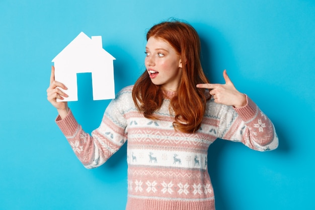 Conceito imobiliário. Mulher ruiva animada com cabelo ruivo, apontando e olhando para o modelo da casa de papel, mostrando o anúncio de apartamento, em pé sobre um fundo azul.