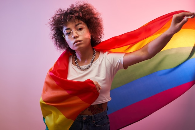 Conceito LGBTQ. Menina caucasiana positiva com cabelo afro encaracolado segurando a bandeira do arco-íris isolada em estúdio