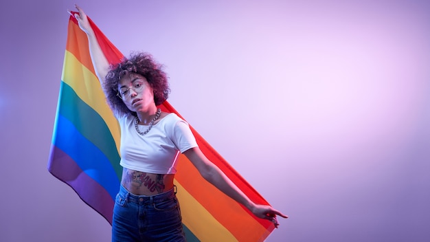 Conceito lgbtq. menina caucasiana positiva com cabelo afro encaracolado segurando a bandeira do arco-íris isolada