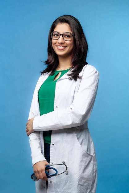 Conceito médico de uma médica asiática linda em um jaleco branco com óculos, na altura da cintura. Estudante de medicina. Uma funcionária do hospital olha para a câmera e sorri, estúdio, com fundo azul