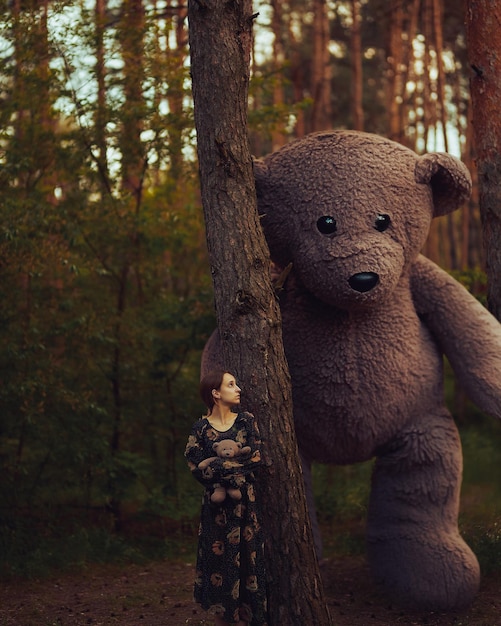 Conceito surrealista - um urso de brinquedo gigante fica ao lado de uma garota em um vestido na floresta