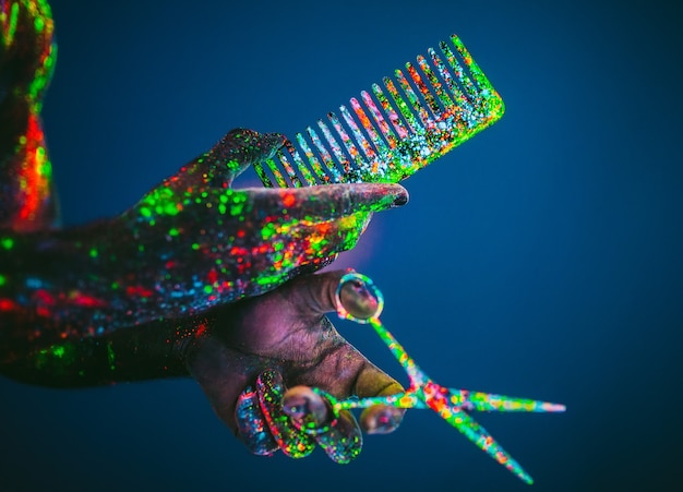 Foto conceito. tesoura e pente são pintados em pó ultravioleta. estilo e moda. mão masculina de barbeiro barbeiro detém tesoura e pente de madeira.