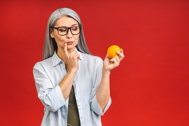 Conceito vegano ou vegetariano Retrato de uma bela idosa asiática madura segurando uma fruta laranja sorrindo isolada sobre fundo vermelho