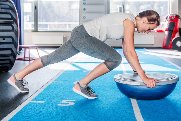 Concentração esportiva linda jovem atlética loira com leggings cinza trabalhando na academia fazendo prancha para os músculos abdominais no treinador de equilíbrio bosu segurando o equilíbrio no fitness ballindoor