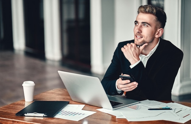 Concentração total. Um jovem empresário está sentado em um centro de negócios com um laptop e um telefone na mão, na mesa estão os documentos e o café