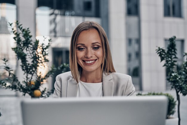 Concentración total en el trabajo. Hermosa joven usando laptop y sonriendo mientras trabaja en interiores