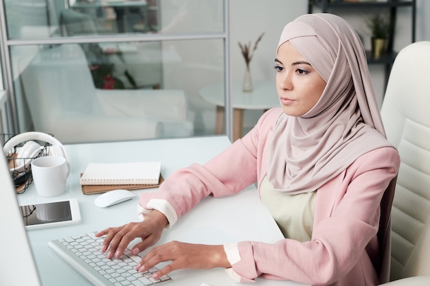 Concentrada jovem empresária muçulmana usando um lenço rosa na cabeça, sentada na mesa e digitando no teclado do computador no escritório
