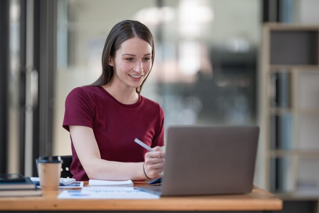 Concentrada en el trabajo Mujer joven segura con ropa casual elegante trabajando en una computadora portátil mientras está sentada cerca de una ventana en una oficina creativa o cafetería