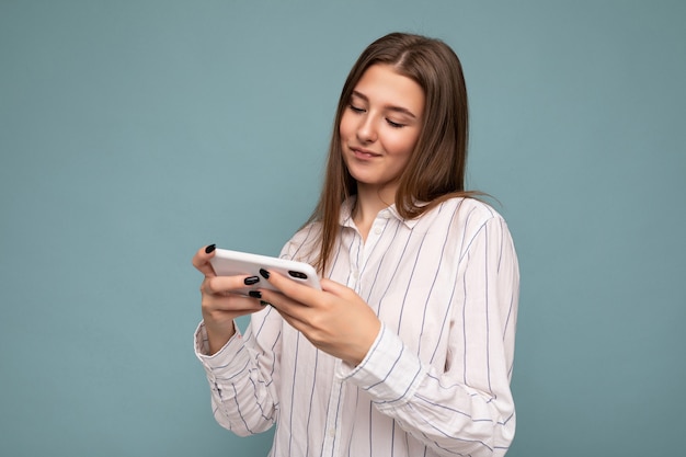 Concentrado feliz atractiva joven rubia vistiendo casual camisa blanca aislada sobre fondo azul pared sosteniendo teléfono inteligente y jugando juegos en línea mirando la pantalla del gadjet.