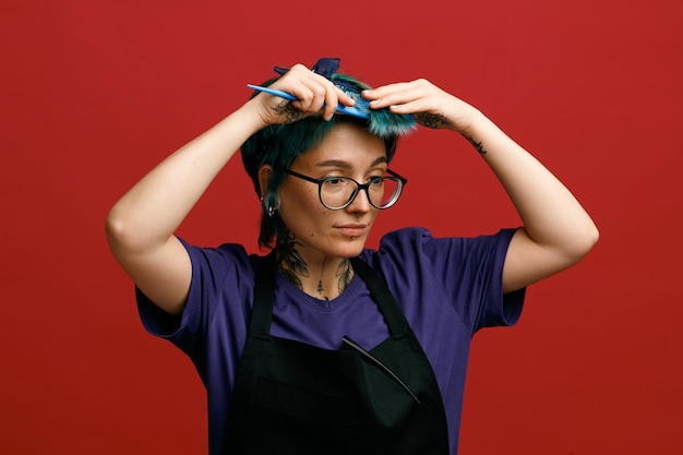 Concentrado jovem barbeiro feminino usando óculos uniformes e bandana olhando para o lado penteando o cabelo com grampo de cabelo no avental isolado em fundo vermelho