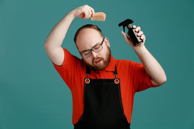 Concentrado jovem barbeiro masculino vestindo óculos camisa vermelha e avental de barbeiro segurando spray de cabelo acima da cabeça e pente perto da cabeça com os olhos fechados isolados no fundo azul